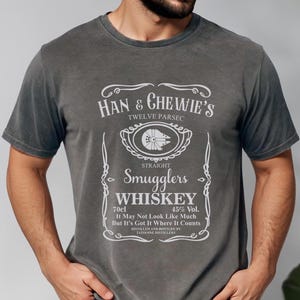 May include: Dark gray t-shirt with a white graphic resembling a whiskey label. The text reads "Han & Chewie's Smugglers Whiskey" with the Millennium Falcon emblem. The shirt also includes the text "70cl" and "45% Vol."