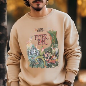 Op de afbeelding: Beige crewneck sweatshirt met een kleurrijke afbeelding van Peter Pan van Walt Disney. De afbeelding toont Peter Pan in een groene outfit, met een zwaard, met andere personages en de tekst "Peter Pan".
