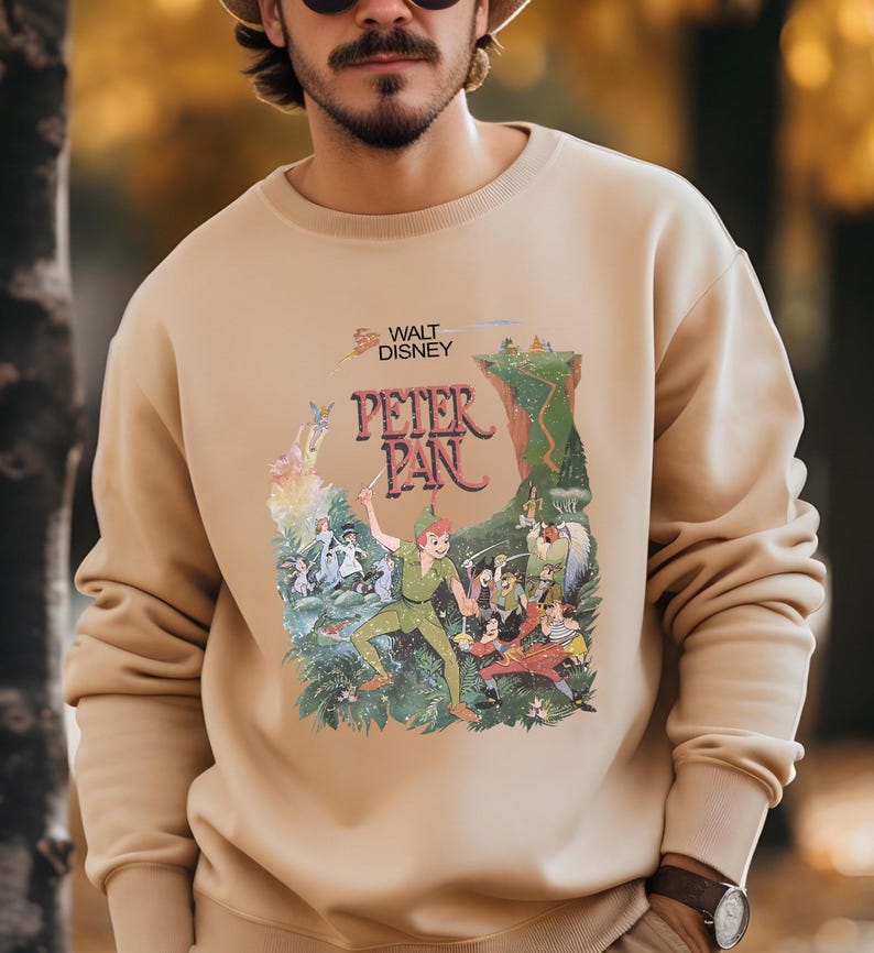 Op de afbeelding: Beige crewneck sweatshirt met een kleurrijke afbeelding van Peter Pan van Walt Disney. De afbeelding toont Peter Pan in een groene outfit, met een zwaard, met andere personages en de tekst "Peter Pan".