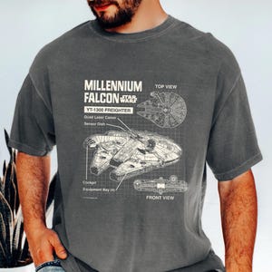 May include: Dark gray t-shirt featuring a detailed blueprint of the Millennium Falcon, a YT-1300 freighter from Star Wars. The design includes top and front views, with labels for key components like the Quad Laser Canon and Sensor Dish.