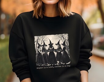May include: Black sweatshirt featuring a vintage-style graphic of five witches in black dresses and hats. The image includes the text "Girls Will Be Girls" and "sugar in a bowl, coffee in a cup."