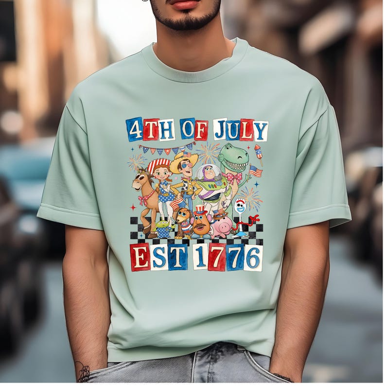 A person wearing a light green t-shirt with a colorful graphic design depicting various cartoon characters and fireworks, celebrating the 4th of July.