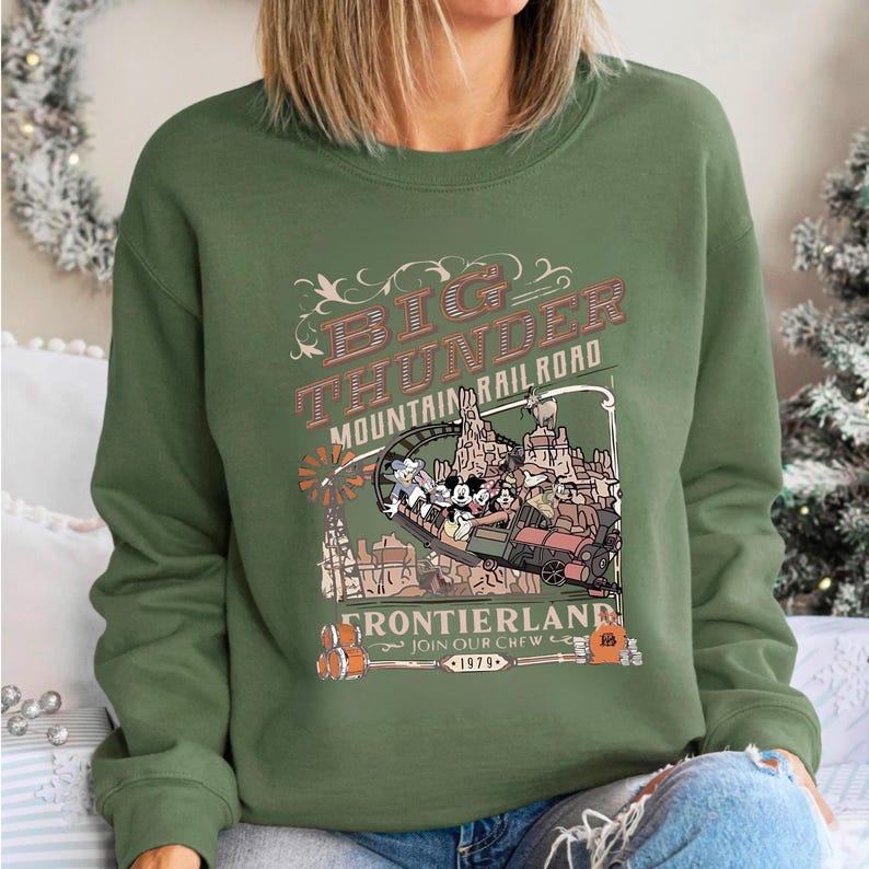 A woman wearing a green sweatshirt with a graphic design depicting a train and a castle, along with the text "BIG THUNDER MOUNTAIN RAILROAD" and "FRONTIERLAND".