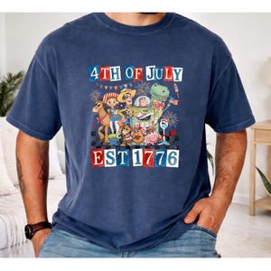 A man is wearing a blue t-shirt with a graphic design depicting cartoon characters and fireworks, celebrating the 4th of July.