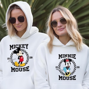 Pu&ograve; includere: Due persone indossano felpe con cappuccio bianche con occhiali da sole. Le felpe presentano grafiche di Topolino e Minnie. La felpa con cappuccio di Topolino ha un cerchio a scacchi in bianco e nero con la scritta "Mickey Mouse" e gli anni "19" e "28". La felpa con cappuccio di Minnie Mouse ha un design simile con la scritta "Minnie Mouse".