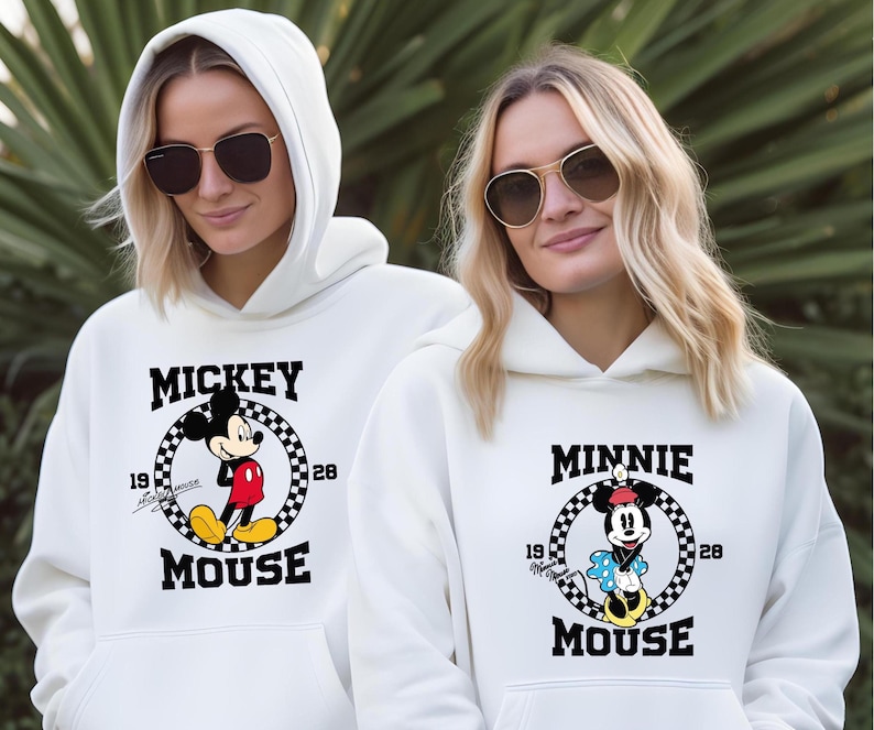 Pu&ograve; includere: Due persone indossano felpe con cappuccio bianche con occhiali da sole. Le felpe presentano grafiche di Topolino e Minnie. La felpa con cappuccio di Topolino ha un cerchio a scacchi in bianco e nero con la scritta "Mickey Mouse" e gli anni "19" e "28". La felpa con cappuccio di Minnie Mouse ha un design simile con la scritta "Minnie Mouse".