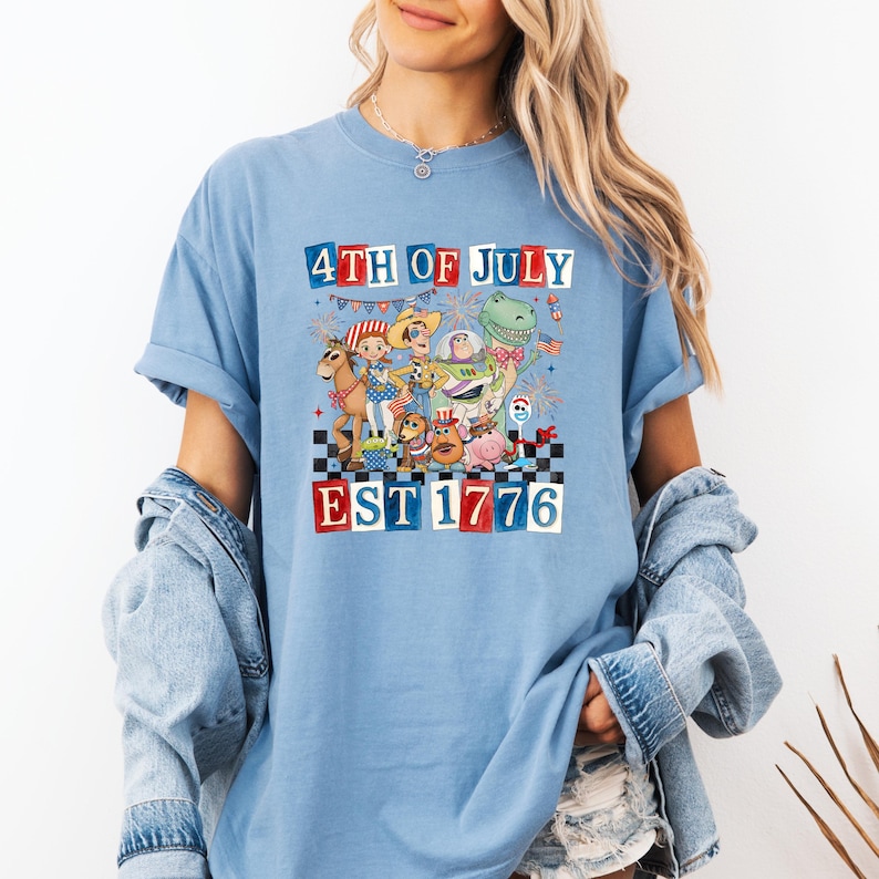 A woman wearing a blue t-shirt with a graphic design depicting cartoon characters and text related to the 4th of July holiday.