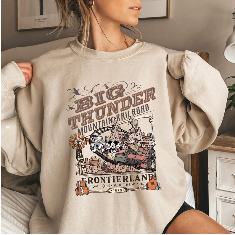 A woman wearing a beige sweatshirt with a graphic design featuring a train and the text "BIG THUNDER MOUNTAIN RAILROAD" and "FRONTIERLAND JOIN OUR CHEW WELCOME".