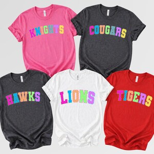 May include: Five short-sleeved t-shirts in pink, dark gray, and red colors. Each shirt features a team name printed in a multi-colored, checkered font across the chest. The names displayed are KNIGHTS, COUGARS, HAWKS, LIONS, and TIGERS.