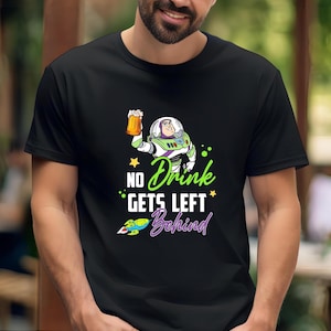 May include: Black t-shirt featuring a cartoon Buzz Lightyear character holding a beer mug. The shirt has the text "No Drink Gets Left Behind" in green and purple, with a rocket ship graphic.
