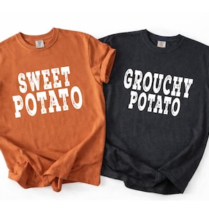 May include: Two short-sleeved crew neck t-shirts. One shirt is burnt orange with the white text "SWEET POTATO" printed on the front. The other shirt is charcoal gray with the white text "GROUCHY POTATO" printed on the front in a matching font.