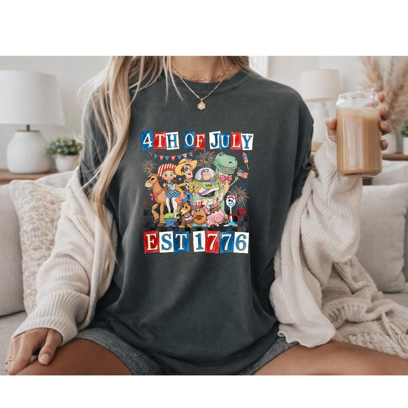 A person wearing a t-shirt with a graphic design celebrating the 4th of July, including a dinosaur, fireworks, and the text "EST 1776".