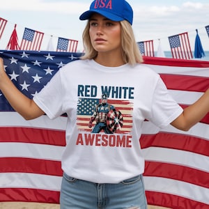 A woman wearing a white t-shirt with a graphic of a superhero and the text "RED WHITE AWESOME" stands in front of an American flag.