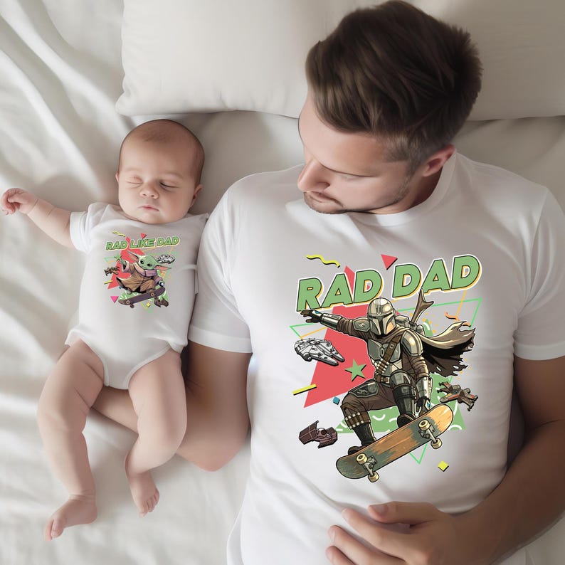 A man and a baby are lying on a bed, with the man wearing a white t-shirt with a graphic of a skateboarder and the baby wearing a white onesie with a graphic of a character from the Star Wars franchise.