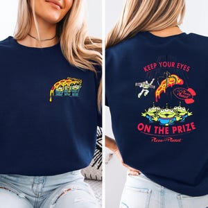 a woman wearing a navy blue t-shirt with a colorful graphic design on the back. The design includes a pizza with various characters from the Toy Story franchise, along with the text "Keep Your Eyes on the Prize" and "Pizza Planet".