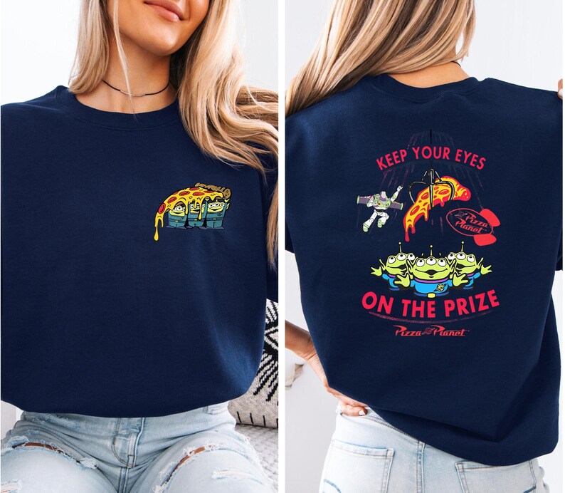 a woman wearing a navy blue t-shirt with a colorful graphic design on the back. The design includes a pizza with various characters from the Toy Story franchise, along with the text "Keep Your Eyes on the Prize" and "Pizza Planet".