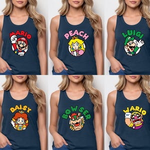 May include: A collection of six navy blue tank tops, each featuring a circular graphic of a Super Mario character. The designs include Mario, Peach, Luigi, Daisy, Bowser, and Wario, with their respective names printed in bold, colorful letters above each character.