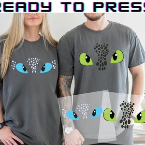 May include: Two gray t-shirts with dragon eye designs. One shirt features blue eyes, the other green. The text "READY TO PRESS" is at the top. A clear transfer sheet with the eye designs is in front of the shirts.