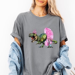 May include: Gray t-shirt featuring a green and blue dinosaur wearing a pink tutu and a pink crown. The dinosaur has its mouth open. The shirt has short sleeves and a relaxed fit.