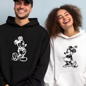Puede incluir: Sudaderas con capucha en blanco y negro con bocetos de Mickey y Minnie Mouse. La sudadera negra tiene a Mickey Mouse, la blanca a Minnie Mouse. Ambas tienen bolsillo delantero y capucha. Ideales para parejas o fans de Disney.