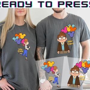 May include: Two gray t-shirts with cartoon illustrations of a man and woman holding Mickey Mouse-shaped balloons. The woman's shirt says "His Ellie," and the man's shirt says "Her Carl."