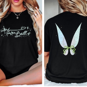 May include: Black t-shirt featuring the text "Tinker Bell" in a whimsical script on the front. The back of the shirt displays a colorful fairy wing design with a green bow. The shirt is a casual, comfortable style.