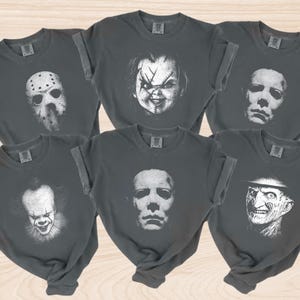 K&ouml;nnte beinhalten: Sechs dunkelgraue T-Shirts mit hochgekrempelten &Auml;rmeln, jeweils mit einem wei&szlig;en Gesicht eines Horrorfilmcharakters. Die Charaktere sind Jason Voorhees, Chucky, Michael Myers, Pennywise und Freddy Krueger.