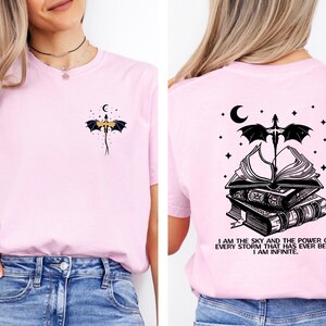 A woman wearing a pink t-shirt with a graphic design on the back. The design features a bat, a crescent moon, and a book, along with the text "I am the sky and the power of every storm that has ever been, I am infinite".