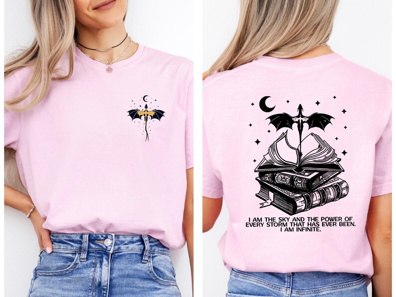 A woman wearing a pink t-shirt with a graphic design on the back. The design features a bat, a crescent moon, and a book, along with the text "I am the sky and the power of every storm that has ever been, I am infinite".