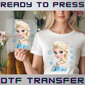 May include: A white t-shirt featuring a cartoon image of a blonde woman with a blue dress and snowflakes. The image is also on a clear transfer sheet held in front of the shirt. The words "READY TO PRESS" and "DTF TRANSFER" are at the top and bottom.