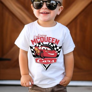 Puede incluir: Camiseta blanca con Lightning McQueen de la pel&iacute;cula Cars. El dise&ntilde;o incluye el texto "Lightning McQueen" encima de un gr&aacute;fico de coche de carreras rojo, con "Cars" debajo. La camiseta tiene un dise&ntilde;o de bandera a cuadros en blanco y negro a cada lado del coche.