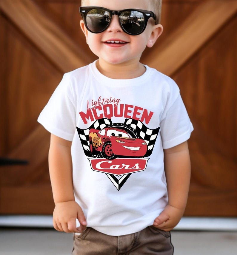 Puede incluir: Camiseta blanca con Lightning McQueen de la pel&iacute;cula Cars. El dise&ntilde;o incluye el texto "Lightning McQueen" encima de un gr&aacute;fico de coche de carreras rojo, con "Cars" debajo. La camiseta tiene un dise&ntilde;o de bandera a cuadros en blanco y negro a cada lado del coche.
