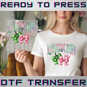 May include: A white t-shirt and a clear transfer sheet featuring the word "WICKED" in a gradient font, with green and pink bows and a crown. The transfer sheet is held up to the shirt, showcasing the design. The words "READY TO PRESS" and "DTF TRANSFER" are also visible.
