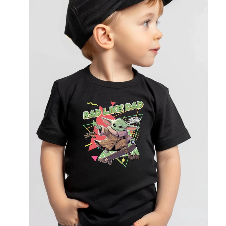 A young boy wearing a black t-shirt with a graphic design featuring a cartoon character riding a skateboard and the text "RAD LIKE DAD" printed on it.