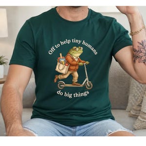 May include: A dark green t-shirt featuring a graphic of a frog wearing glasses and a brown cardigan, riding a scooter while carrying a tote bag filled with school supplies. The text reads, Off to help tiny humans do big things.