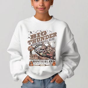 A young woman wearing a white sweatshirt with a graphic design featuring a cartoon-style illustration of a train and a cityscape.