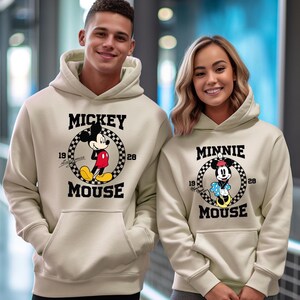 Pu&ograve; includere: Due felpe con cappuccio beige con Topolino e Minnie Mouse. La felpa con cappuccio di Topolino ha un cerchio a scacchi in bianco e nero con Topolino al centro, il testo "MICKEY MOUSE" e i numeri "19" e "28". La felpa con cappuccio di Minnie Mouse ha un design simile.