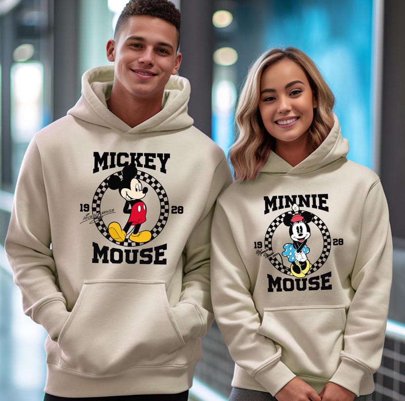 Pu&ograve; includere: Due felpe con cappuccio beige con Topolino e Minnie Mouse. La felpa con cappuccio di Topolino ha un cerchio a scacchi in bianco e nero con Topolino al centro, il testo "MICKEY MOUSE" e i numeri "19" e "28". La felpa con cappuccio di Minnie Mouse ha un design simile.