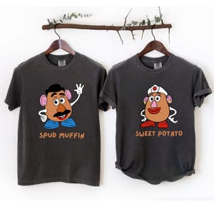 May include: Two dark gray t-shirts with cartoon potato head designs. One shirt says "Spud Muffin" and features a potato head with a mustache and blue shoes. The other says "Sweet Potato" and has a potato head wearing a white hat.