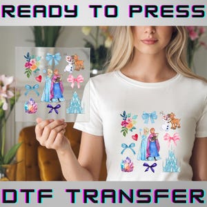 May include: A clear DTF transfer sheet featuring watercolor illustrations of characters, bows, and a castle. The text "READY TO PRESS" and "DTF TRANSFER" are displayed in a bold, colorful font. The transfer is held up against a white t-shirt with a matching design.