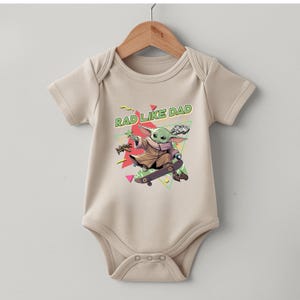 A beige baby onesie with a graphic design featuring a cartoon character riding a skateboard and the text "RAD LIKE DAD" printed on it.