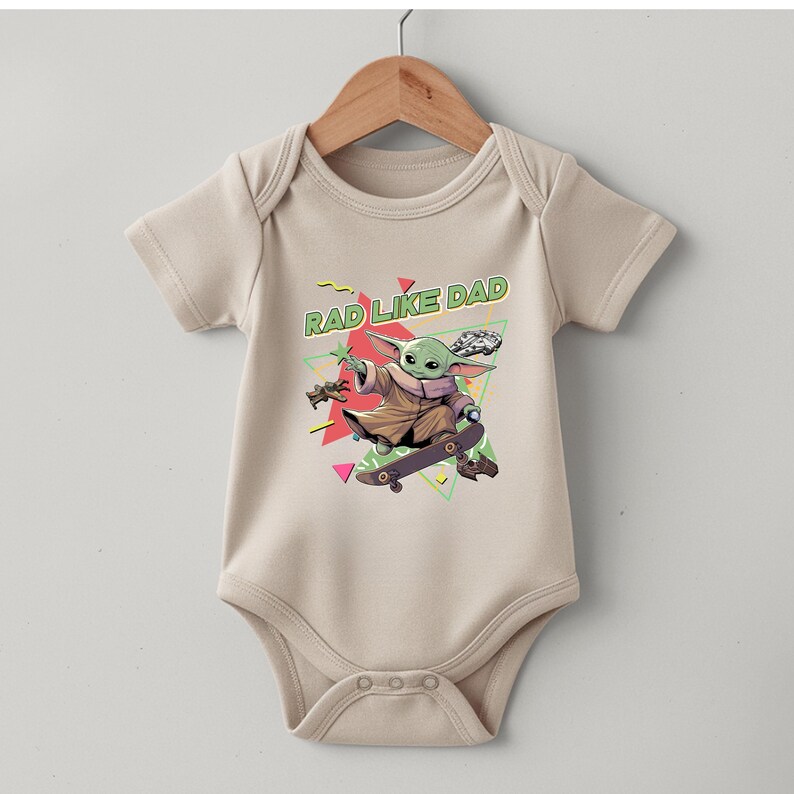 A beige baby onesie with a graphic design featuring a cartoon character riding a skateboard and the text "RAD LIKE DAD" printed on it.