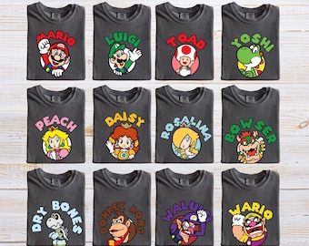 May include: Twelve dark gray t-shirts arranged in a grid, each featuring a circular graphic of a Nintendo character. Characters include Mario, Luigi, Toad, Yoshi, Peach, Daisy, Rosalina, Bowser, Dry Bones, Donkey Kong, Waluigi, and Wario with names.