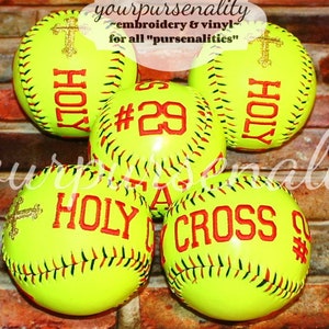 May include: Five bright yellow softballs featuring red embroidered text and cross designs. The balls display the words HOLY, CROSS, and the number 29. A watermark with the text yourpursenality embroidery & vinyl for all pursenalities is visible.