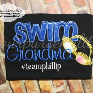 Może przedstawiać: Czarna koszulka z wyhaftowanym napisem SWIM z niebieskiego brokatu, Grandma niebieską nicią oraz #teamphillip białą nicią. Po prawej stronie napisu znajduje się haftowana grafika ż&oacute;łtych okular&oacute;w pływackich z opalizującymi szkłami.