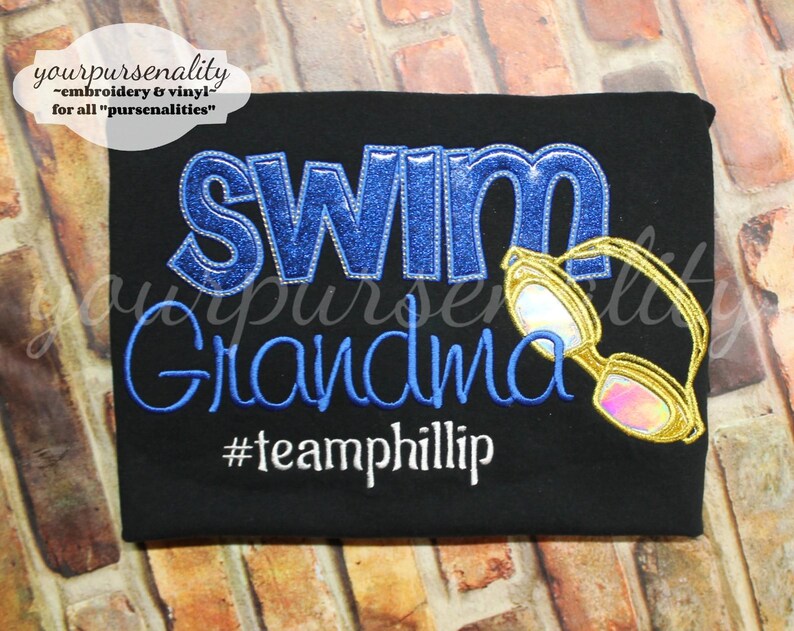 Może przedstawiać: Czarna koszulka z wyhaftowanym napisem SWIM z niebieskiego brokatu, Grandma niebieską nicią oraz #teamphillip białą nicią. Po prawej stronie napisu znajduje się haftowana grafika ż&oacute;łtych okular&oacute;w pływackich z opalizującymi szkłami.
