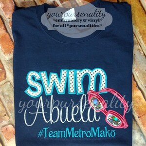 Może przedstawiać: Granatowa koszulka z wyhaftowanym napisem SWIM w turkusowe kropki na białym tle, słowem Abuela białą kursywą oraz #TeamMetroMako w kolorze turkusowym. Po prawej stronie wyhaftowano r&oacute;żowe okulary pływackie z opalizującymi szkłami.