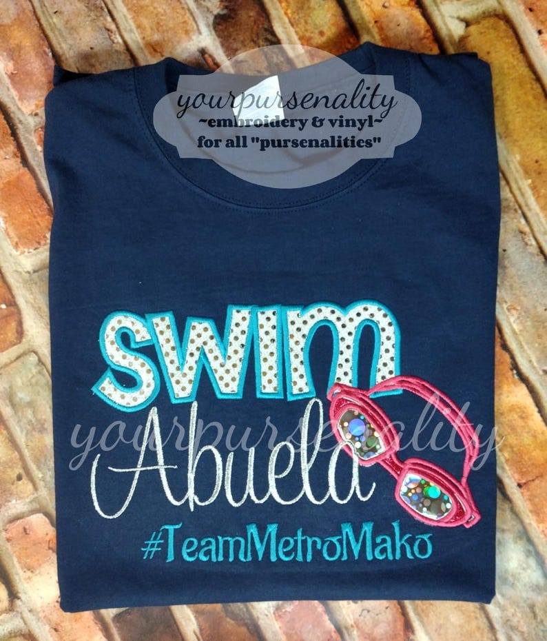 Może przedstawiać: Granatowa koszulka z wyhaftowanym napisem SWIM w turkusowe kropki na białym tle, słowem Abuela białą kursywą oraz #TeamMetroMako w kolorze turkusowym. Po prawej stronie wyhaftowano r&oacute;żowe okulary pływackie z opalizującymi szkłami.
