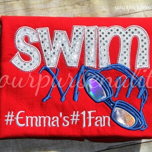 Może przedstawiać: Jaskrawoczerwony t-shirt z napisem SWIM wykonanym ze srebrnego materiału w kropki, słowem Mom napisanym niebieską kursywą oraz białym haftowanym hashtagiem #Emma's#1Fan. Dołączono niebieski haftowany wz&oacute;r okular&oacute;w pływackich z opalizującymi szkłami.
