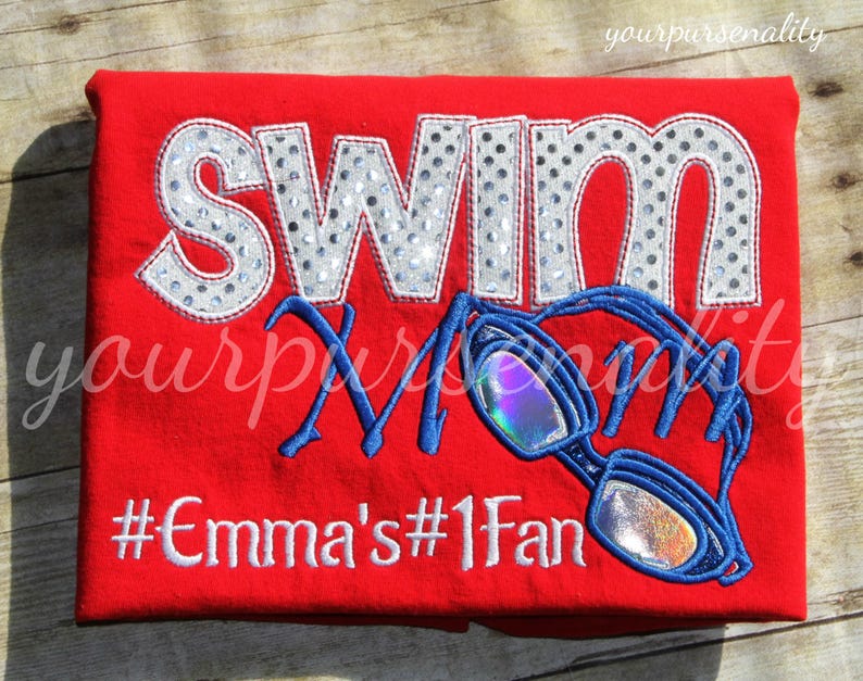 Może przedstawiać: Jaskrawoczerwony t-shirt z napisem SWIM wykonanym ze srebrnego materiału w kropki, słowem Mom napisanym niebieską kursywą oraz białym haftowanym hashtagiem #Emma's#1Fan. Dołączono niebieski haftowany wz&oacute;r okular&oacute;w pływackich z opalizującymi szkłami.